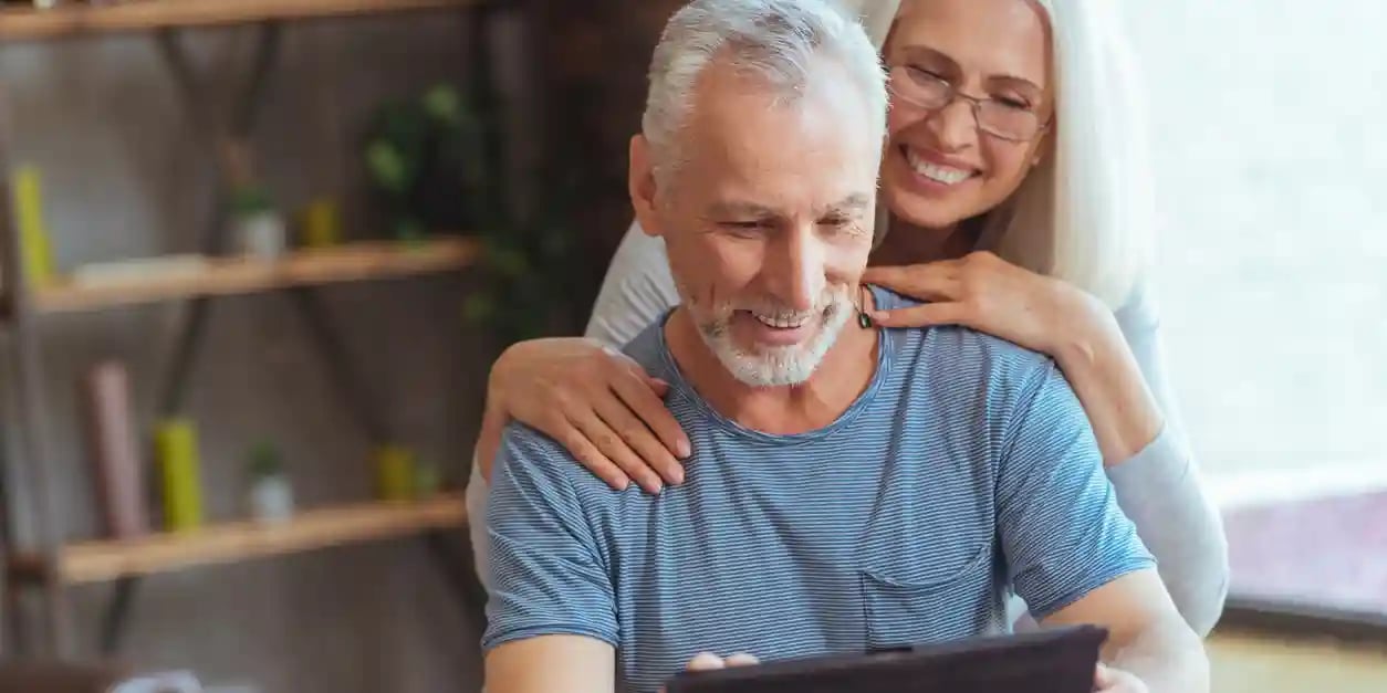 A retired man using a tablet and sitting at the table while resting with his wife at home.