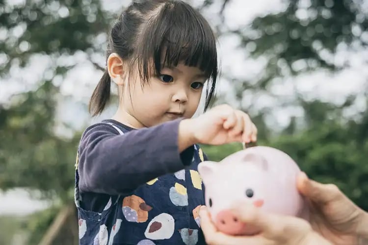 A parent out of the shot teaching her toddler about money by giving her a piggy bank.